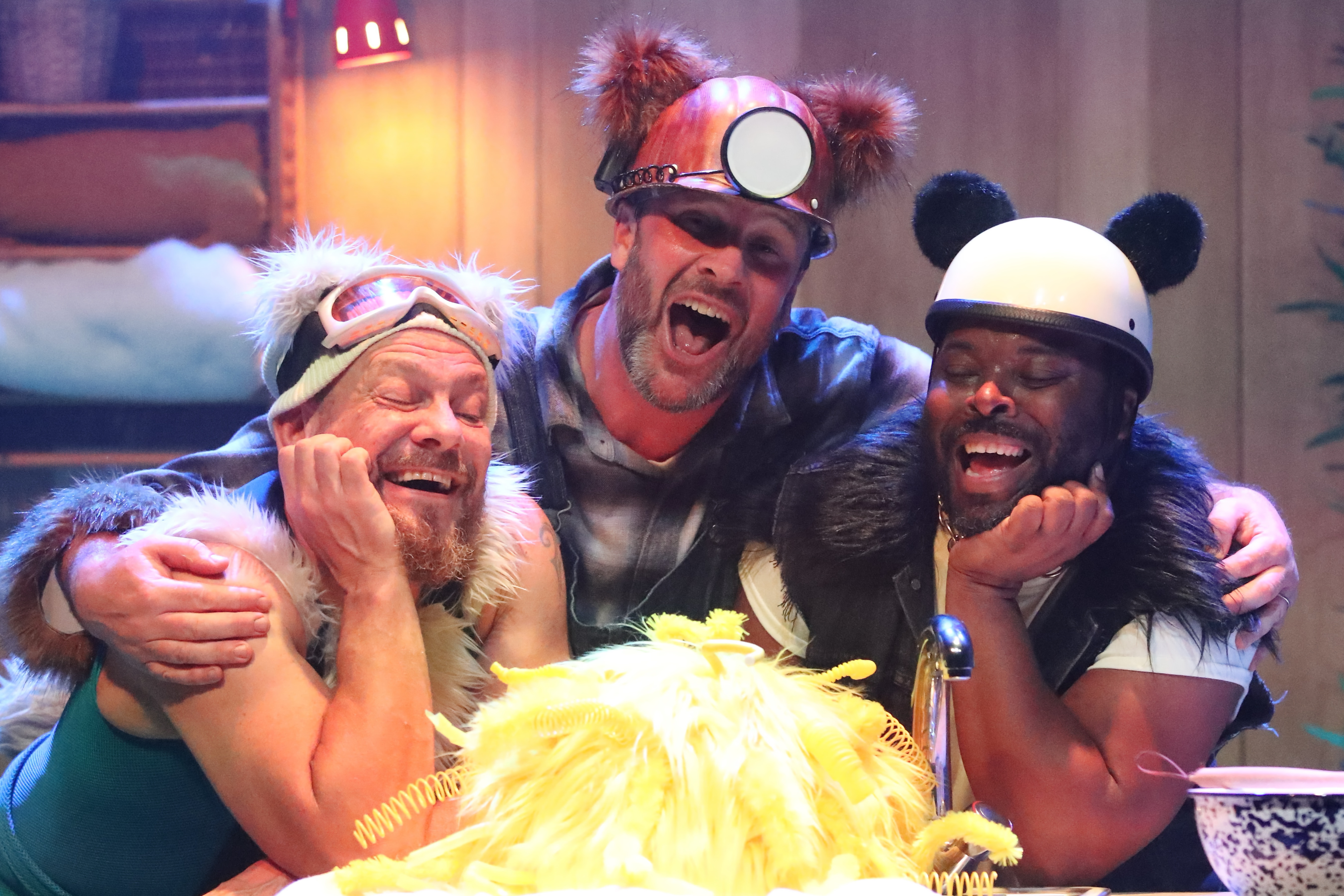 A stage scene from The Three Bears. Three performers are behind a kitchen cabinet. Panda, a bear inspired by a panda, is wearing black and white clothes with a black fur trim. Polar, a bear inspired by a polar bear, is wearing a blue ski suit, ski hat and goggles with white fur trim. Grizzly, a bear inspired by a grizzly bear, wears dungarees and a miner's hat with brown fur trim. They all look lovingly at ‘Goldie’ a mass of bright yellow feathers who is sat infant of them.
